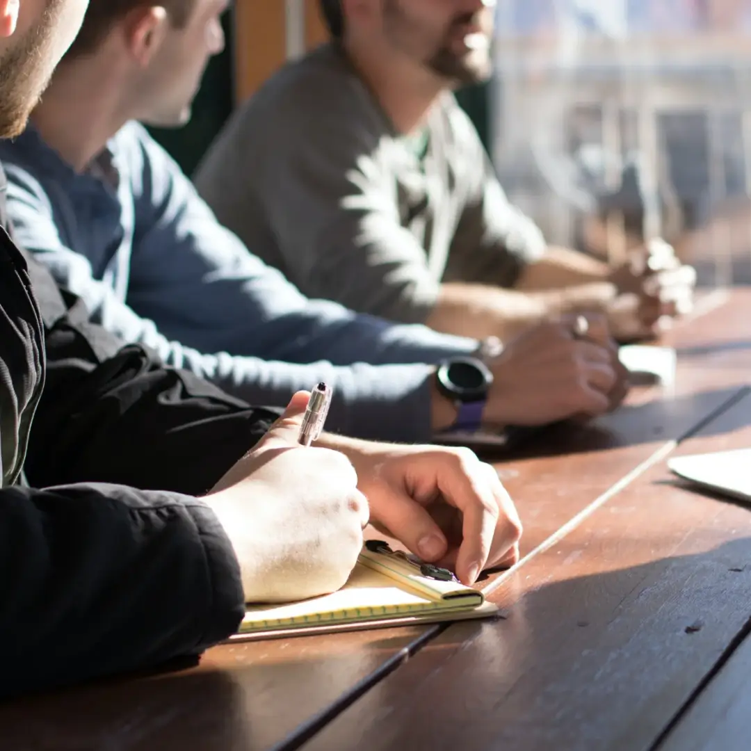 People taking notes during a meeting at a wooden table.
